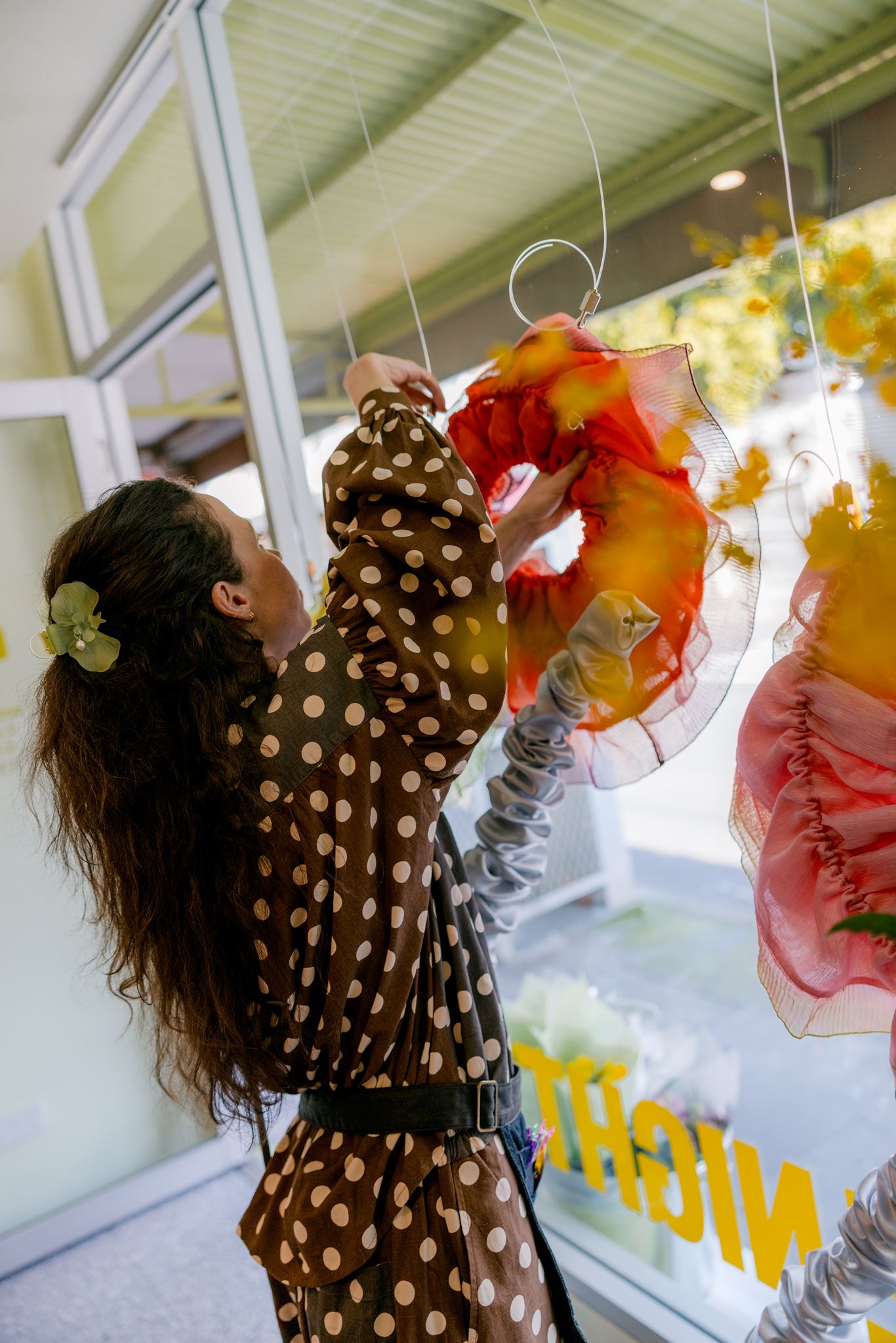 A woman in a brown and beige polka dot dress hangs a colorful orange and yellow fabric flower display in front of a glass window.